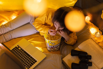 Wall Mural - little 11 year old girl using laptop under her home-made tent inside the living room and eating popc