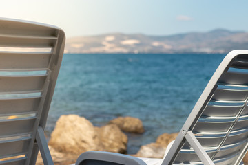 View of two white foldable benches near the sea, concept of vacation and summer. Bright blue water in the background