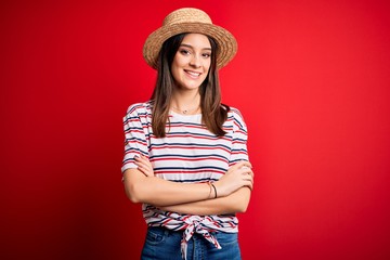 Young beautiful brunette woman wearing striped t-shirt and summer hat over red background happy face smiling with crossed arms looking at the camera. Positive person.