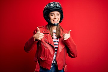 Wall Mural - Young beautiful brunette motocyclist woman wearing motorcycle helmet and red jacket success sign doing positive gesture with hand, thumbs up smiling and happy. Cheerful expression and winner gesture.