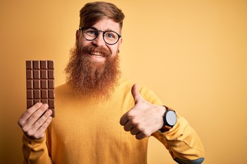 Irish redhead man with beard eating a sweet chocolate bar over yellow isolated background happy with big smile doing ok sign, thumb up with fingers, excellent sign