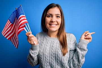 Canvas Print - Young patriotic woman holding usa flag on independence day 4th of july over blue background very happy pointing with hand and finger to the side