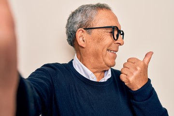 Canvas Print - Senior handsome grey-haired man wearing sweater and glasses making selfie by camera pointing and showing with thumb up to the side with happy face smiling