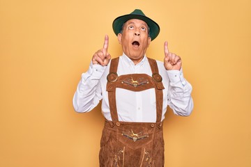 Canvas Print - Senior grey-haired man wearing german traditional octoberfest suit over yellow background amazed and surprised looking up and pointing with fingers and raised arms.