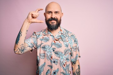 Poster - Handsome bald man with beard and tattoo wearing casual floral shirt over pink background smiling and confident gesturing with hand doing small size sign with fingers looking and the camera. Measure