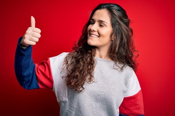 Sticker - Young beautiful woman with curly hair wearing casual sweatshirt over isolated red background Looking proud, smiling doing thumbs up gesture to the side