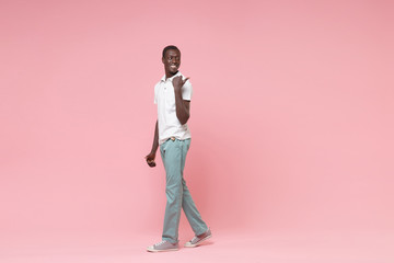 Side view of smiling young african american man guy in white polo shirt, turquoise trousers posing isolated on pastel pink background. People lifestyle concept. Mock up copy space. Point thumb aside.