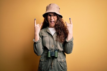 Poster - Young beautiful tourist woman on vacation wearing explorer hat and binoculars shouting with crazy expression doing rock symbol with hands up. Music star. Heavy concept.