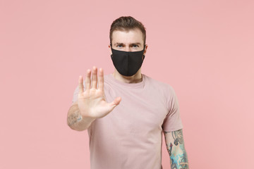 Young tattooed man guy in pastel casual t-shirt, black face mask posing isolated on pink wall background studio portrait. People lifestyle concept. Mock up copy space. Showing stop gesture with palm.