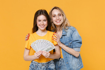 Two smiling young women girls friends in casual t-shirts denim clothes isolated on yellow background studio. People lifestyle concept. Mock up copy space. Hold fan of cash money in dollar banknotes.