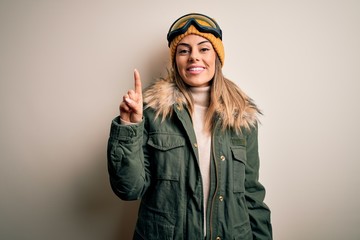 Sticker - Young brunette skier woman wearing snow clothes and ski goggles over white background pointing finger up with successful idea. Exited and happy. Number one.