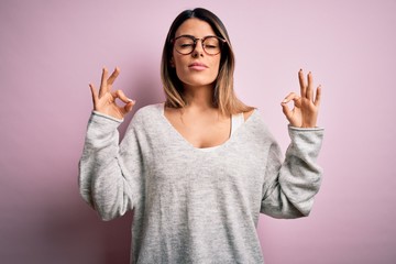 Wall Mural - Young beautiful brunette woman wearing casual sweater and glasses over pink background relax and smiling with eyes closed doing meditation gesture with fingers. Yoga concept.