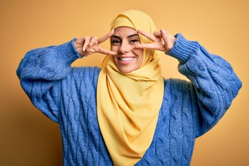 Sticker - Young beautiful brunette muslim woman wearing arab hijab over isolated yellow background Doing peace symbol with fingers over face, smiling cheerful showing victory