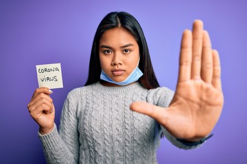 Asian girl wearing medical mask asking for alert holding reminder with corona virus message with open hand doing stop sign with serious and confident expression, defense gesture