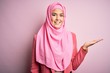 © Krakenimages.com - Young beautiful girl wearing muslim hijab standing over isolated pink background smiling cheerful presenting and pointing with palm of hand looking at the camera.