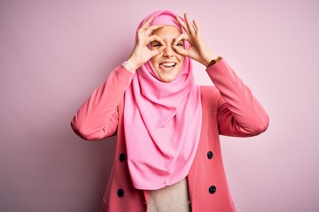 Poster - Young beautiful girl wearing muslim hijab standing over isolated pink background doing ok gesture like binoculars sticking tongue out, eyes looking through fingers. Crazy expression.