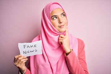 Canvas Print - Young beautiful girl wearing muslim hijab holding paper with fake news message serious face thinking about question, very confused idea