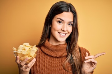 Poster - Young beautiful girl holding bowl with chips potatoes standing over yellow background very happy pointing with hand and finger to the side