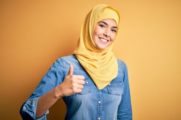 Canvas Print - Young beautiful girl wearing muslim hijab standing over isolated yellow background doing happy thumbs up gesture with hand. Approving expression looking at the camera showing success.