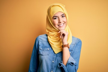 Wall Mural - Young beautiful girl wearing muslim hijab standing over isolated yellow background looking confident at the camera with smile with crossed arms and hand raised on chin. Thinking positive.