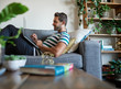 © mavoimages - Smiling man using a laptop while sitting on his sofa