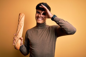 Poster - Young handsome man holding homemade fresh bread over isolated yellow background with happy face smiling doing ok sign with hand on eye looking through fingers