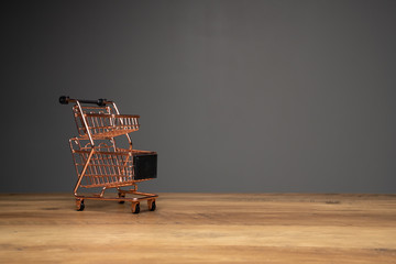 Small empty shopping cart or metal trolley in copper colour on top of wooden top and dark background with copy space.