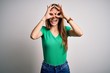 © Krakenimages.com - Young beautiful redhead woman wearing casual green t-shirt and glasses over white background doing ok gesture like binoculars sticking tongue out, eyes looking through fingers. Crazy expression.