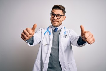 Young doctor man with blue eyes wearing medical coat and stethoscope over isolated background approving doing positive gesture with hand, thumbs up smiling and happy for success. Winner gesture.