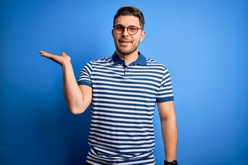 Young man with blue eyes wearing glasses and casual striped t-shirt over blue background smiling cheerful presenting and pointing with palm of hand looking at the camera.