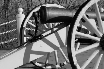 Black and white highly contrasted partial shot of front of an old cannon with two large wheels and fence in background