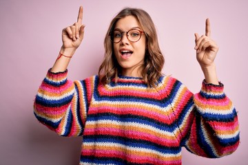 Young beautiful blonde girl wearing glasses and casual sweater over pink isolated background smiling amazed and surprised and pointing up with fingers and raised arms.