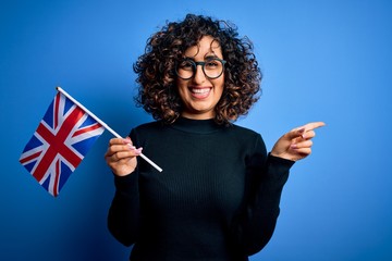 Canvas Print - Young beautiful curly arab woman wearing glasses holding uk flag over blue background very happy pointing with hand and finger to the side