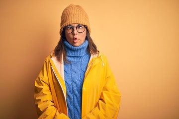 Sticker - Middle age woman wearing yellow raincoat and winter hat over isolated background making fish face with lips, crazy and comical gesture. Funny expression.