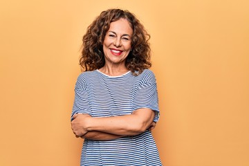 Middle age beautiful woman wearing striped t-shirt standing over isolated yellow background happy face smiling with crossed arms looking at the camera. Positive person.