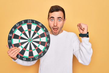 Wall Mural - Young handsome man with blue eyes holding dartboard over isolated yellow background screaming proud and celebrating victory and success very excited, cheering emotion