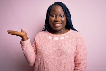 Sticker - African american plus size woman with braids wearing casual sweater over pink background smiling cheerful presenting and pointing with palm of hand looking at the camera.