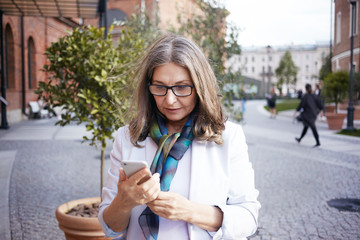 Wall Mural - Portrait of busy modern middle aged European woman in stylish clothes standing on street, checking time on mobile, having nervous look, being late for business meeting. Gadgets and technology concept