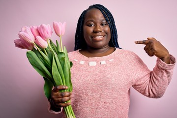 Sticker - Young african american plus size woman with braids holding bouquet of pink tulips flower looking confident with smile on face, pointing oneself with fingers proud and happy.