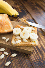 Wall Mural - Toasts with peanut paste and banana on the cutting board on the brown wooden background. Location vertical. Closeup.