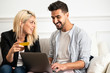 © Antonio Ovejero/ADDICTIVE STOCK - Delighted blonde female smiling and reading credit card credentials to cheerful ethnic boyfriend with laptop while sitting on sofa and making online purchases together