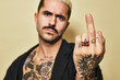 © Cien X Cien Studio/ADDICTIVE STOCK - Young aggressive provocative male in black coat over naked tattooed torso showing middle finger gesture while standing against beige background