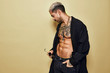 © Cien X Cien Studio/ADDICTIVE STOCK - Brutal muscular sexy fit male with tattooed torso wearing black coat and trendy jeans with stylish sunglasses and accessories standing against beige background looking away