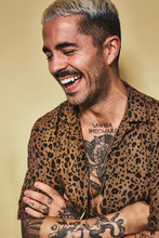 Portrait Of Cheerful Fashionable Male Model With Tattoos Wearing Trendy Leopard Shirt Standing Against Beige Background With Closed Eyes