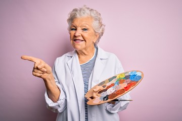 Senior beautiful grey-haired artist woman painting using brush and palette over pink background very happy pointing with hand and finger to the side