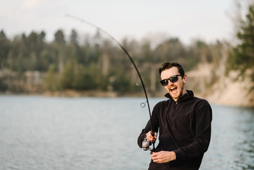 Wall Mural - Fisherman with rod, spinning reel on the river bank. Man catching fish, pulling rod while fishing from lake or pond with text space at weekend. Fishing for pike, perch, carp on beach lake or pond.