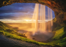 Majestic Waterfall Located On Entrance Of Grassy Stone Cave Against Cloudy Sundown Sky On Seashore In Iceland