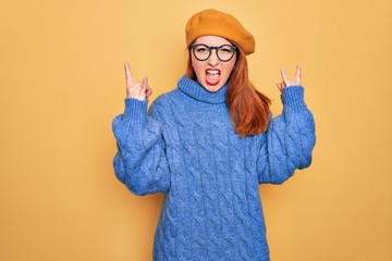 Canvas Print - Young beautiful redhead woman wearing french beret and glasses over yellow background shouting with crazy expression doing rock symbol with hands up. Music star. Heavy concept.