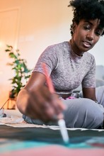 Young Beautiful African American Woman With Afro Hair, Amateur Artist In Casual Outfit Drawing Colorful Picture With Paintbrush On Paper While Sitting On Floor In Cozy Apartment