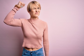 Sticker - Young blonde woman with short hair wearing casual turtleneck sweater over pink background angry and mad raising fist frustrated and furious while shouting with anger. Rage and aggressive concept.